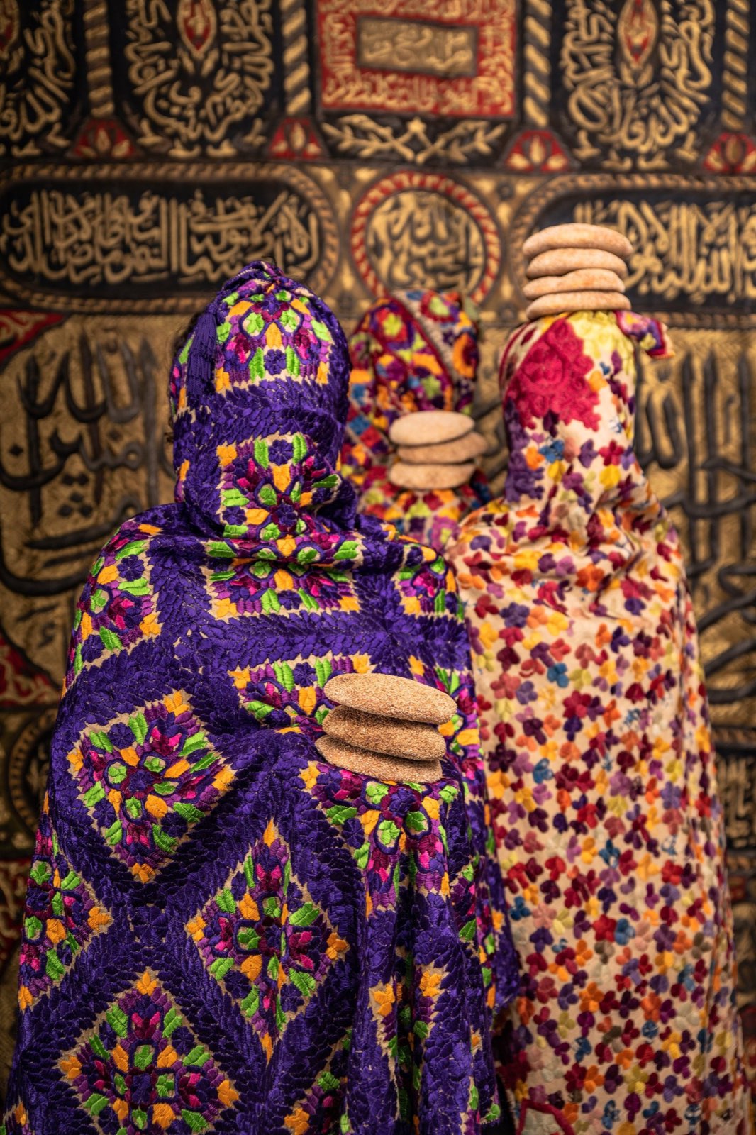 Three figures draped in vivid embroidered cloth before gold Kaaba calligraphy, stones balanced on their heads.