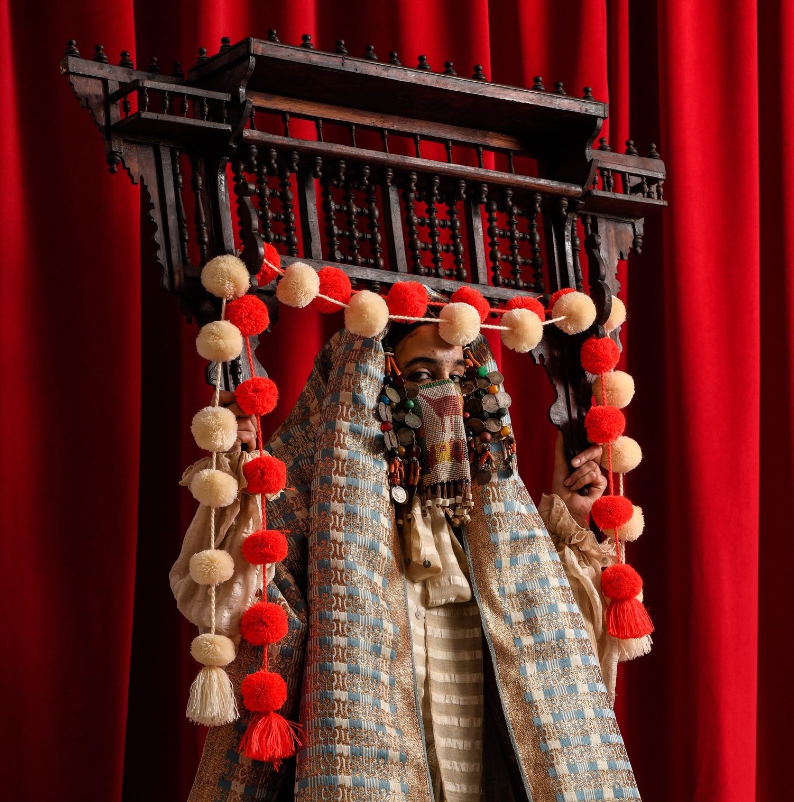 The Mashrabiya Veil — a latticed cedar window strung with wedding pompoms, held before a veiled face.