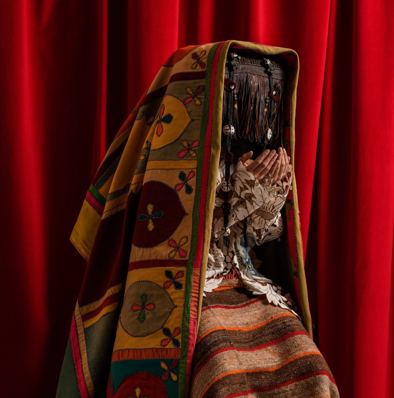 A Maison ARTC figure draped in Berber appliqué textile, hands raised, against a red curtain.