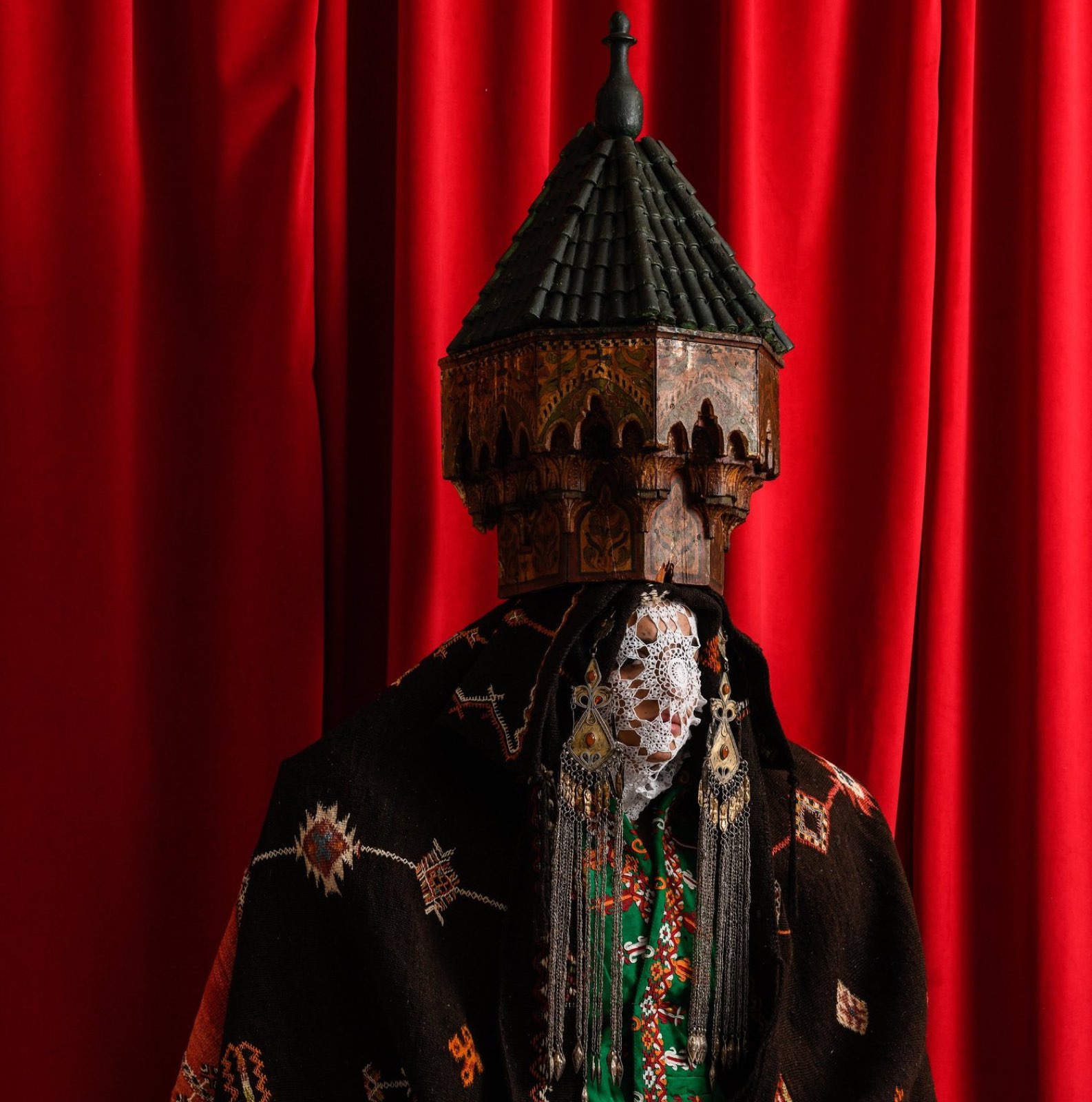 A Maison ARTC piece — an antique carved Moroccan minaret worn as a headdress over a lace veil and kilim, against red.