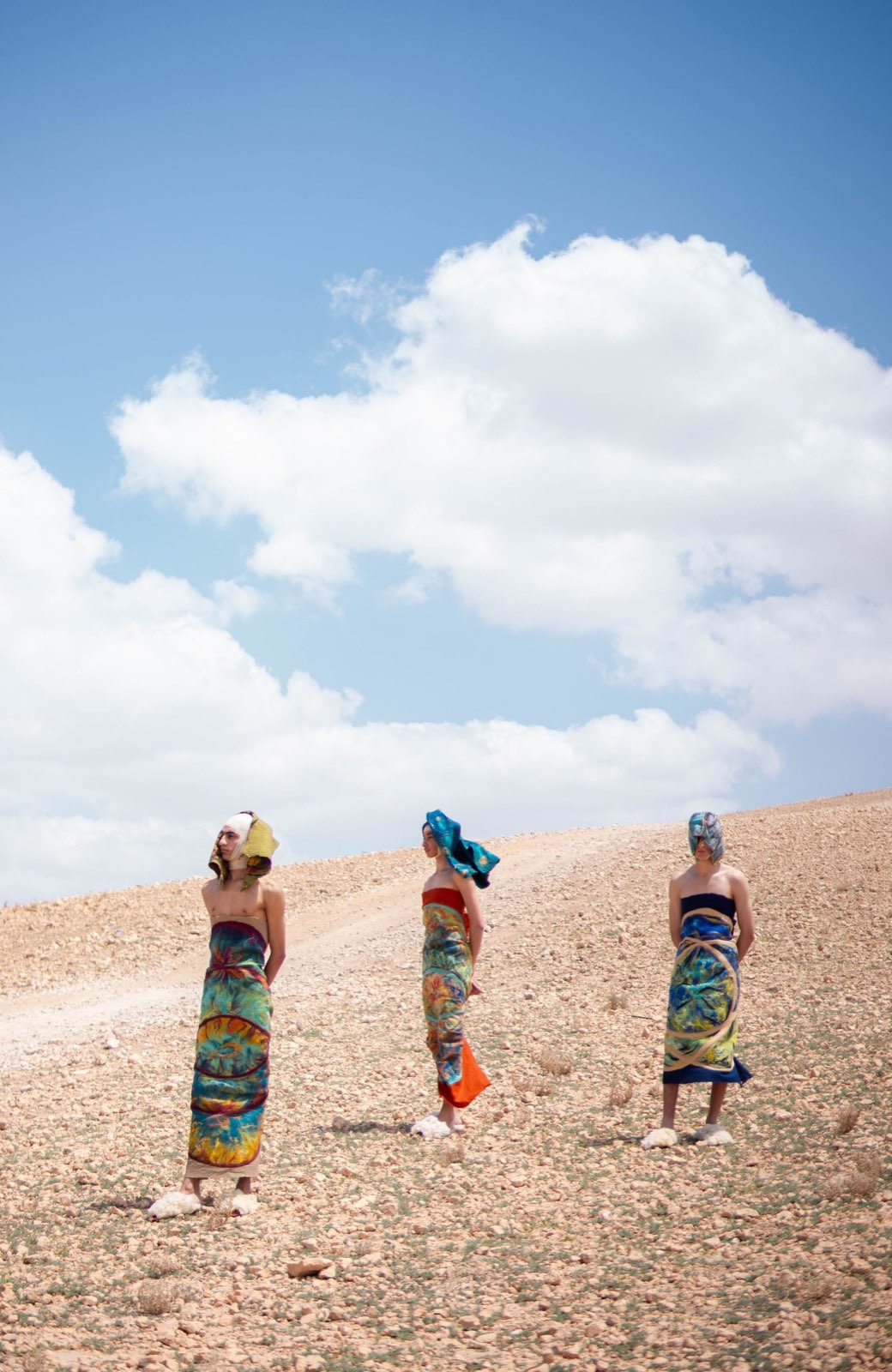 Three figures in painted felted Maison ARTC wraps standing on a Moroccan hillside beneath a wide sky.