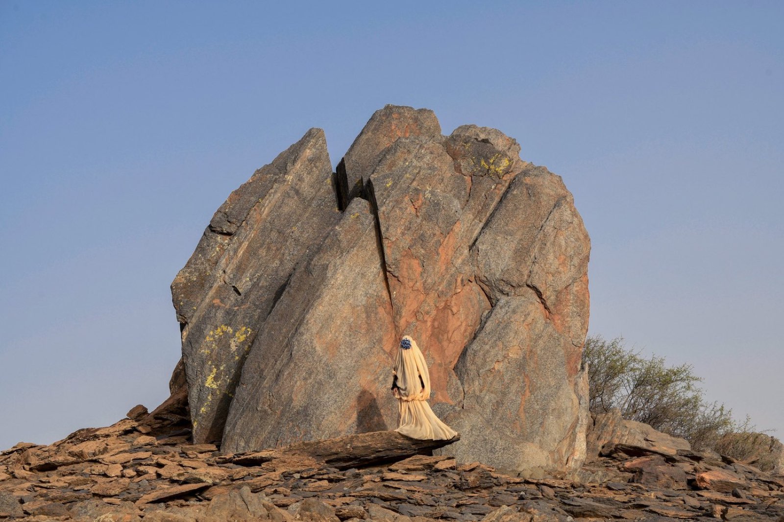 A lone veiled figure in a Maison ARTC piece walking beneath a great split boulder in the Moroccan desert.
