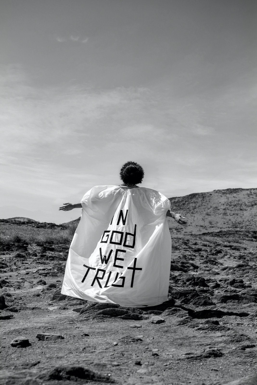A figure in a white cape reading In God We Trust, in the desert — black and white.